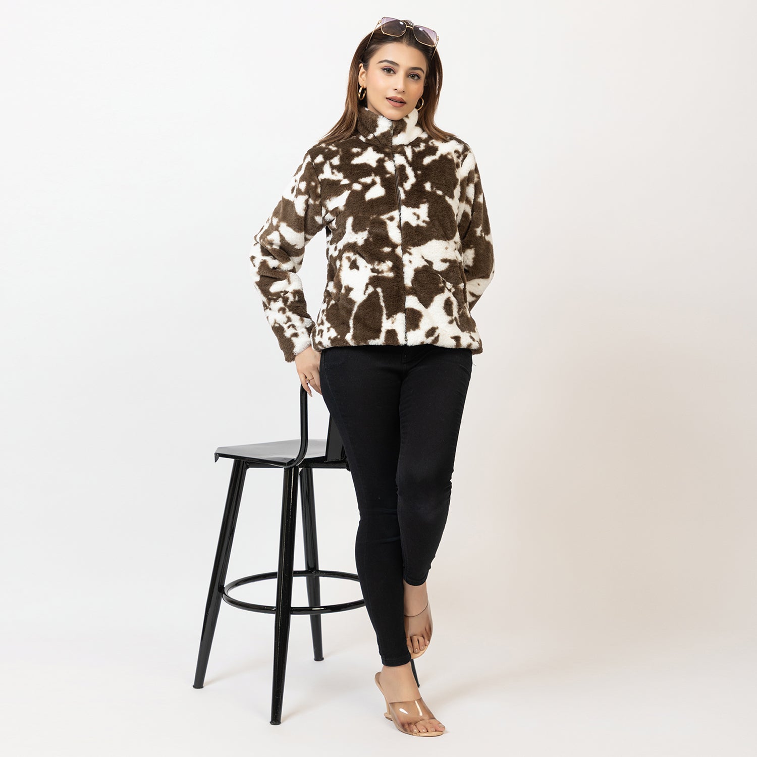 Woman wearing a patterned sweater and black pants standing next to a black stool on a white background Amoldo